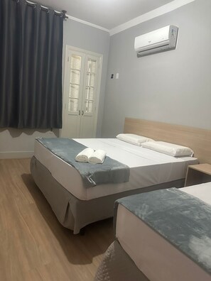 A bedroom with a bed, a nightstand, an air conditioning unit, a window with curtains, and a door with shutters. - Unity Hotel - Santos (Santos)