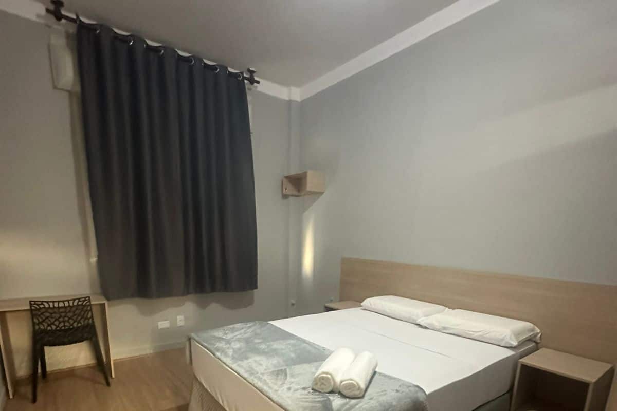 A neatly arranged hotel room with a bed, a small bedside table, a chair, and a window with curtains.