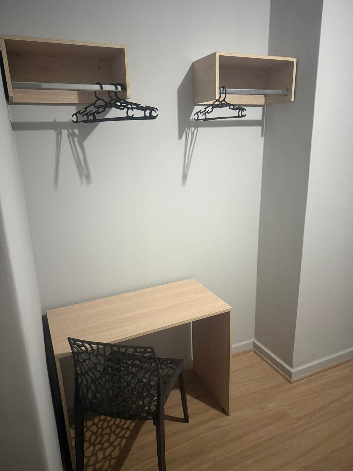 Habitación de esquina con un escritorio de madera, una silla negra y dos estanterías de madera con perchas negras.