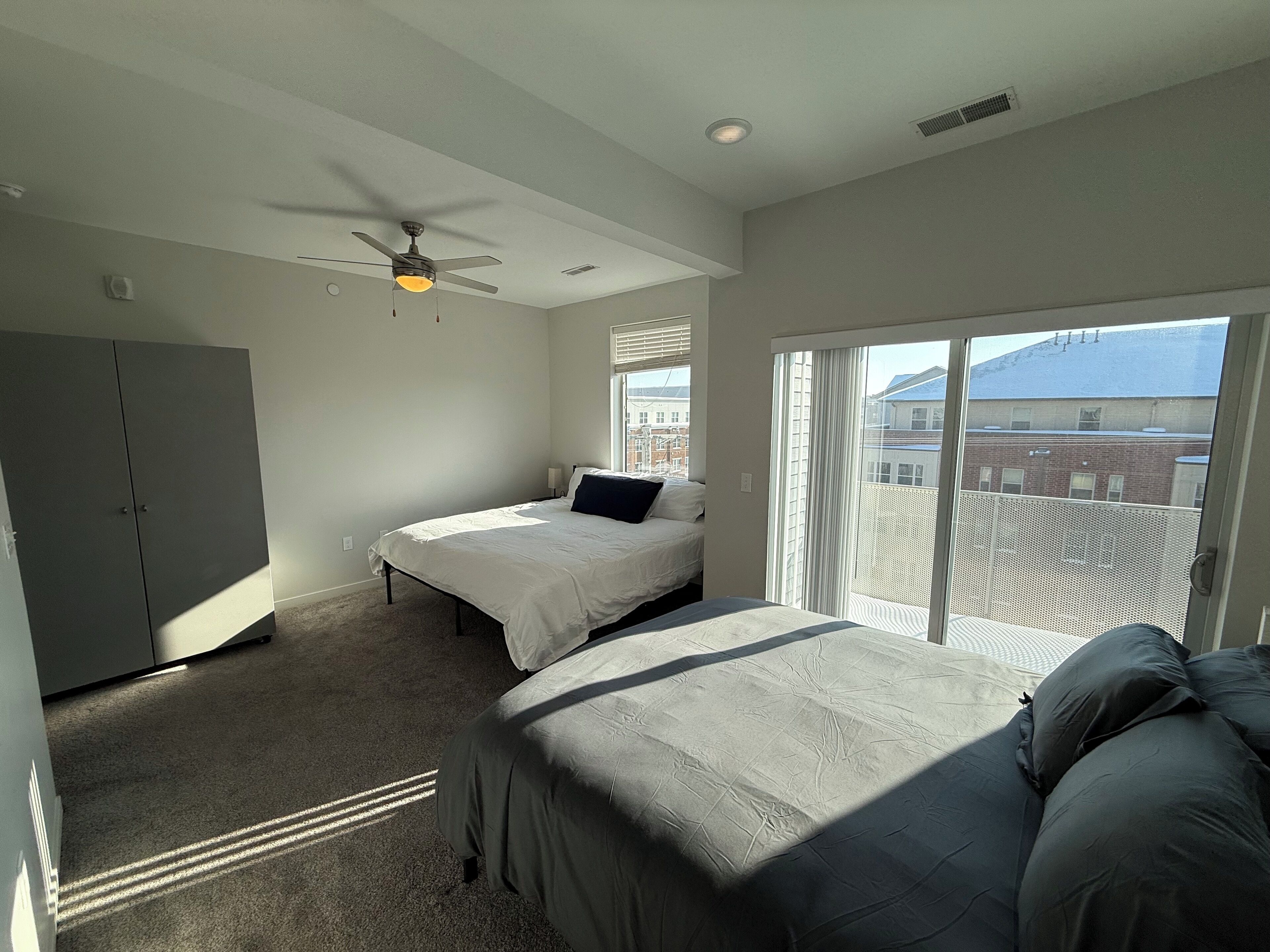 A bedroom with two beds, a ceiling fan, and a large window with a view of buildings.
