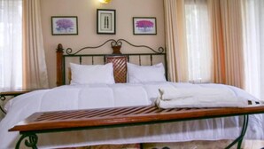 Una cama bien hecha con sábanas blancas, cabecera de madera y estructura metálica. Hay dos cuadros enmarcados en la pared sobre la cama.
