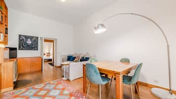 Ein modernes Wohnzimmer mit einem Holzes Esstisch, türkisfarbenen Stühlen, einem gemusterten Teppich, einer Stehlampe und einer Couch mit Kissen.