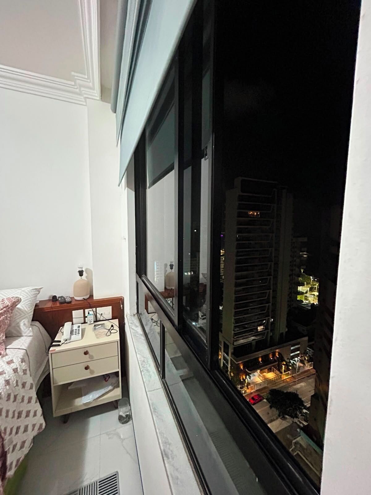 Un balcón con cama, mesita de noche y vista a la ciudad iluminada de noche.