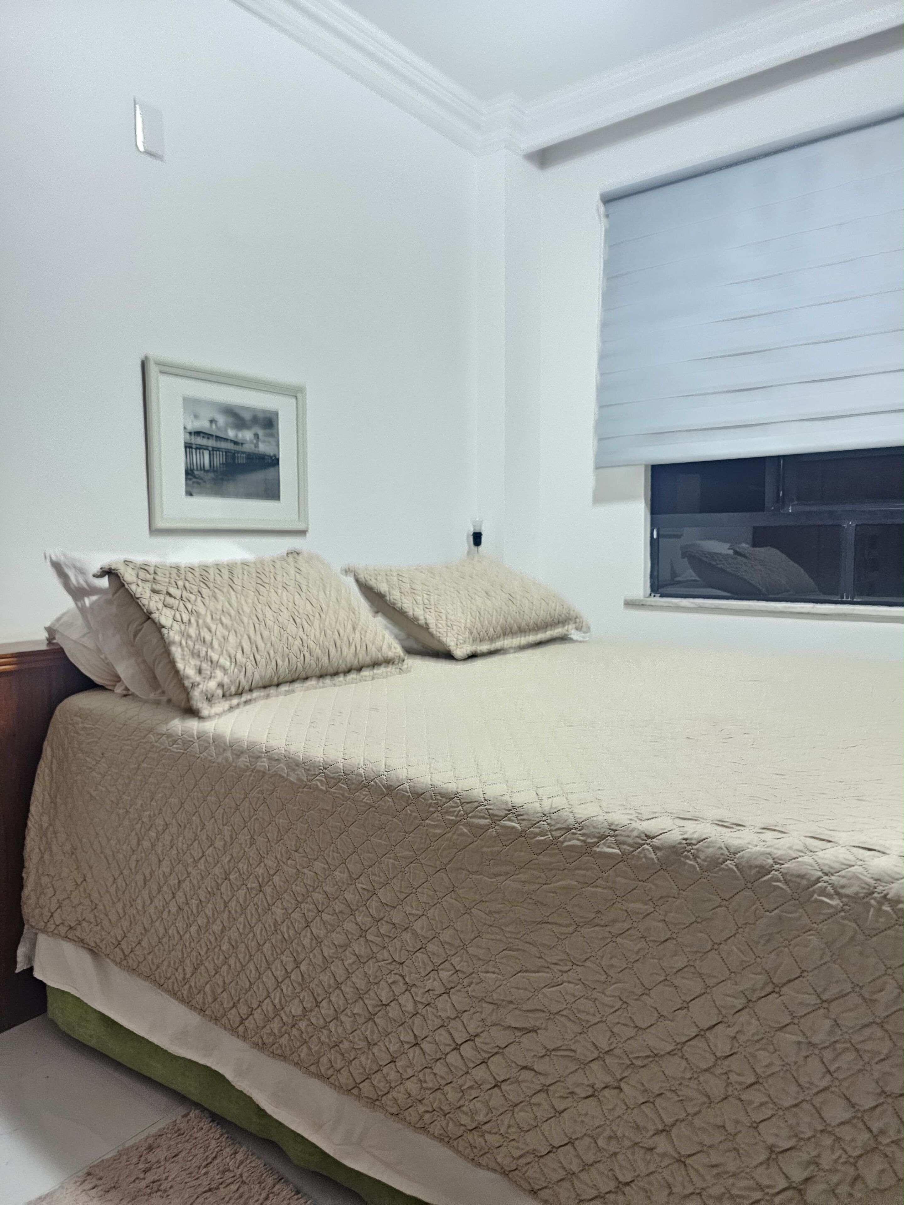 Un dormitorio con cama, almohadas, un cuadro en la pared y una ventana con persianas.