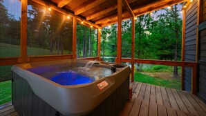 A hot tub on a wooden deck with string lights, surrounded by a wooden deck and a view of a forest. - Double Magic - Unique cabin for 6 - pond, hot tub (Broken Bow)