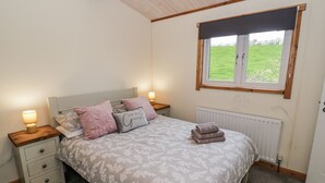 A bedroom with a bed, two bedside tables, a nightstand, a window with a view of the outdoors, and a radiator. - Willow Lodge (Rugeley)