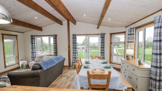 A dining area with a wooden table set for four, surrounded by chairs. There is a sofa with a blanket and a small table with a lamp. Large windows with curtains provide views of the outdoors. - Willow Lodge (Rugeley)
