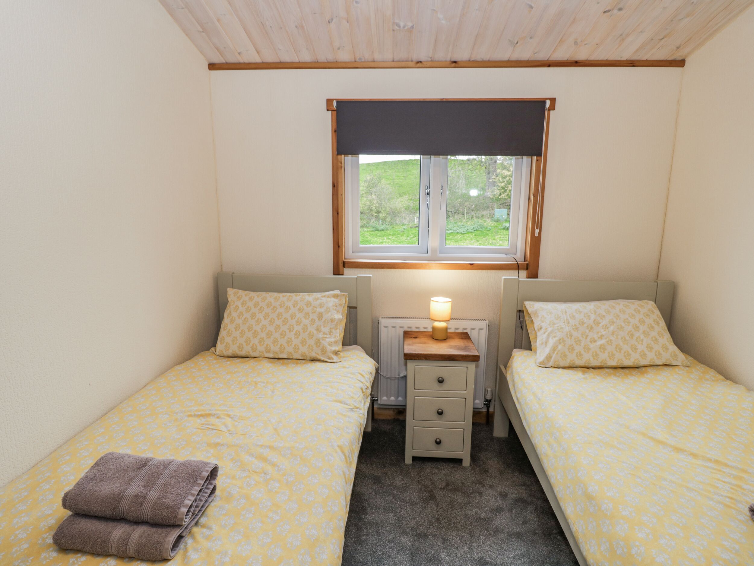 Un dormitorio pequeño y acogedor con dos camas individuales, una mesita de noche con lámpara, una ventana con vistas a la vegetación y un radiador.