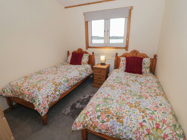 A bedroom with two single beds, a nightstand with a lamp, a window with a view, and a wooden headboard. - Oak Lodge (Rugeley)