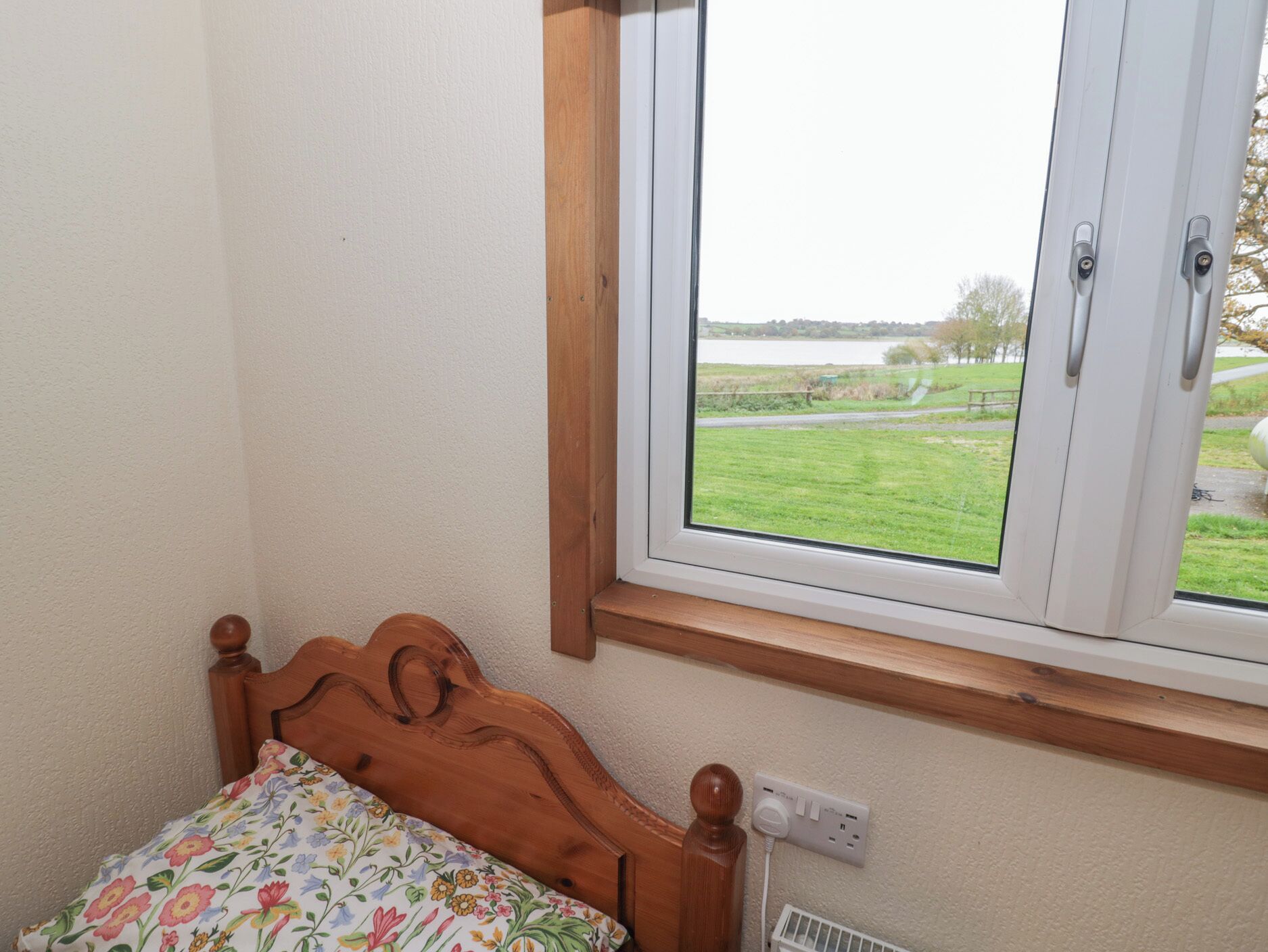 A bedroom with a wooden bed, a window with a view of a grassy field and a body of water, and a wall socket.
