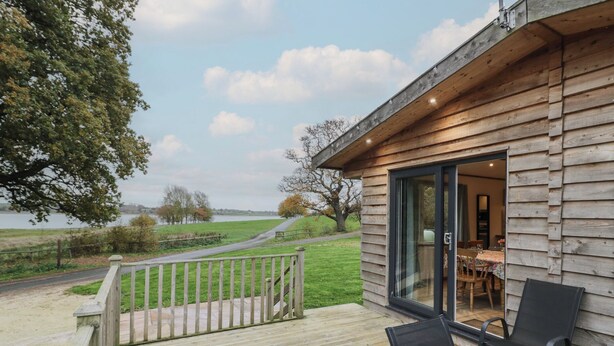 En træhytte med terrasse, spiseplads med stole og udsigt over en sø.
