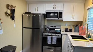 A modern kitchen with a stainless steel refrigerator, microwave, stove, and oven. There is a trash bin and a window with blinds. - Earthy Luxe Guesthouse-Fully Fenced- W/D in unit (Hesperia)