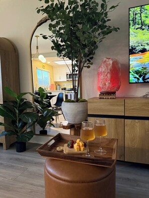Una sala de estar acogedora con una mesa de centro de madera que sostiene una bandeja con aperitivos y bebidas, una planta grande en maceta y un televisor que muestra una escena de naturaleza.