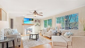 A spacious living room with a fireplace, large windows, and a ceiling fan.