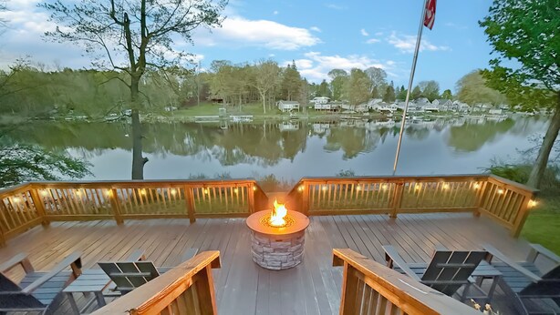 A wooden deck with a central fire pit, surrounded by benches and railings, overlooking a calm lake with houses in the background.
