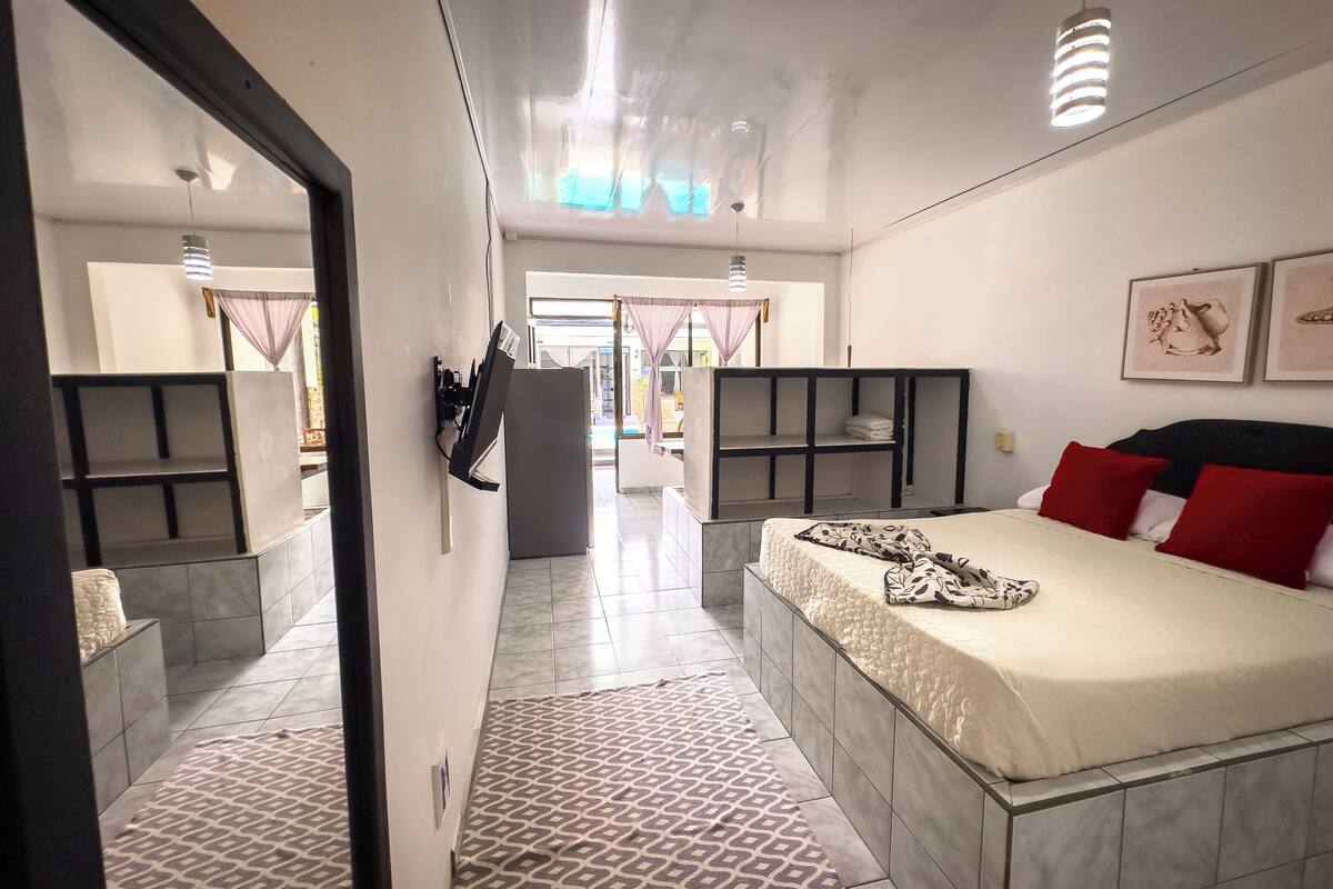 A modern hotel room with a large bed, a desk, and a wardrobe. There is a view of the hallway and a balcony.