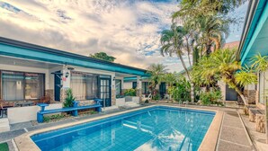 A pool area with a clear blue pool, a covered seating area with chairs, and surrounding tropical plants. - Ela Kosher Hotel and Jewish Center (Jaco)
