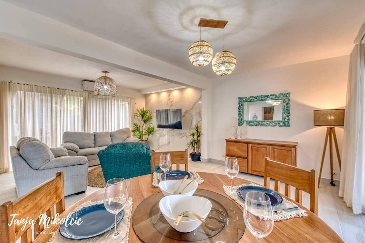 A dining area with a wooden table set for four, a grey sofa, and a teal armchair in a modern living space with a marble wall and decorative mirror.