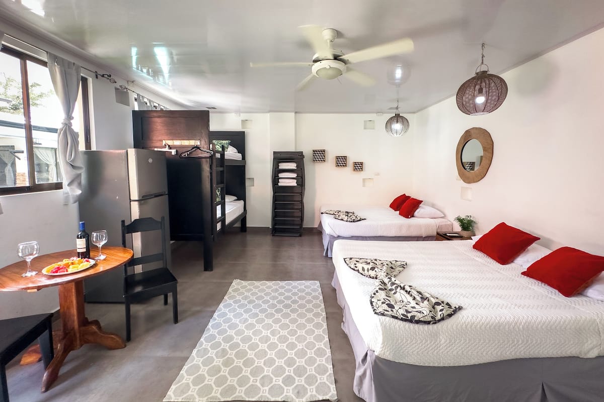 A compact hotel room with two beds, a dining area, a kitchenette, and a ceiling fan.