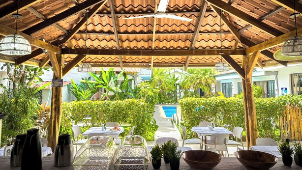 A covered outdoor seating area with tables and chairs, surrounded by lush greenery and a view of a swimming pool. - Ela Kosher Hotel and Jewish Center (Jaco)