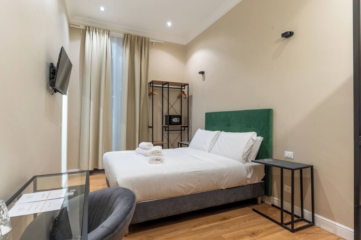 A hotel room with a bed, a desk with papers, a chair, a TV mounted on the wall, and a window with curtains.