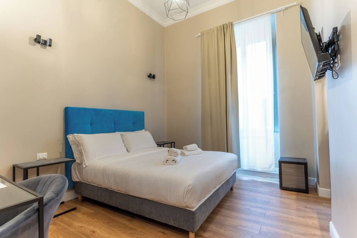 A bedroom with a bed, a desk, a chair, a small table, a wall-mounted TV, and a window with curtains.