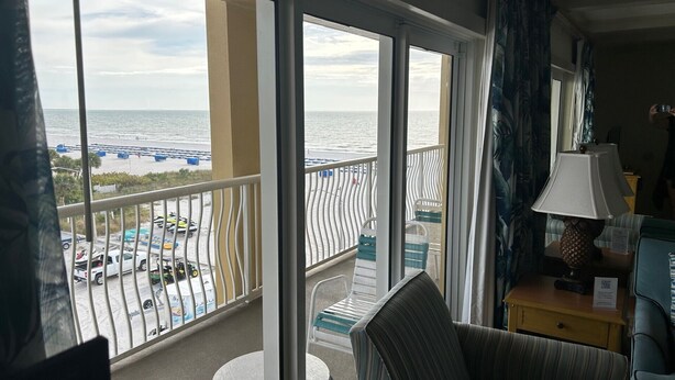 En balkon med udsigt over havet og stranden, et lille bord, en stol og en lampe.