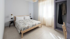 Une chambre avec un lit en bois, deux tables de chevet avec des lampes, une fenêtre avec des rideaux légers et un portant à vêtements avec des vestes.