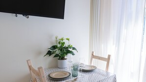 Une table à manger est dressée avec des assiettes, des couverts et un verre d’eau. On y voit également une plante en pot et un téléviseur fixé au mur.