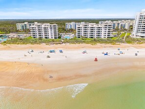 A beach with multiple buildings in the background, a clear sky, and a sandy shore. - Shorehom by the Sea 63 (New Smyrna Beach)