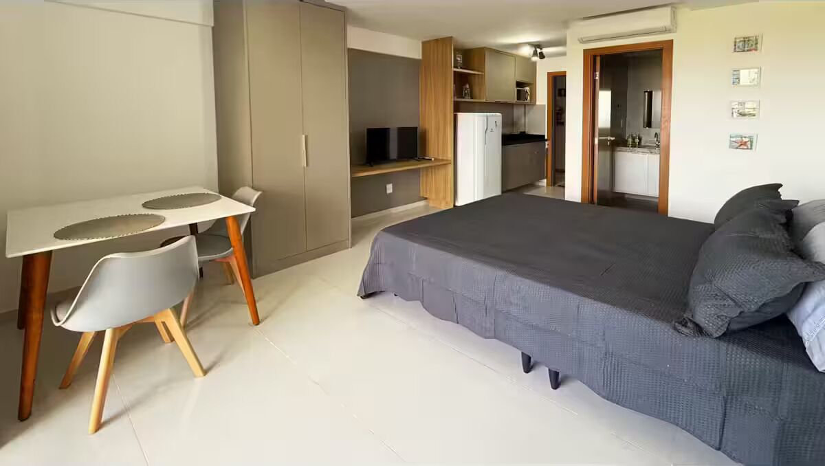 Ein Schlafzimmer mit einem Bett, einem Schreibtisch und einem Stuhl. Im Hintergrund ist eine Küchenzeile zu sehen.