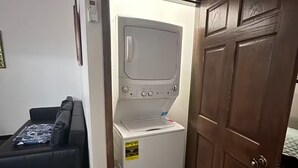A compact laundry room with a washing machine and dryer, a wooden door, and a black leather sofa with a patterned throw pillow.