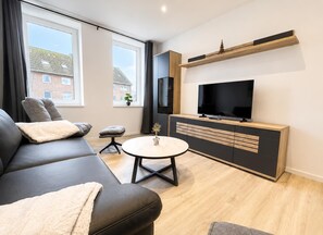 A modern living room with a sofa, ottoman, coffee table, TV, and built-in shelving. - House Deichlust Whg.2,Strichweg 165, 27476 Cuxhaven-Döse (Cuxhaven-Döse)