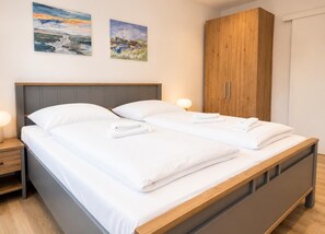 A double bed with white linens and two pillows, a wooden headboard, a wooden wardrobe, and two paintings on the wall.