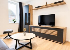A modern living room with a TV on a wooden cabinet, a round table with a vase, a grey sofa, and a window with curtains. - Deichlust House, Apt. 2, Strichweg 165, 27476 Cuxhaven-Döse (Cuxhaven-Döse)