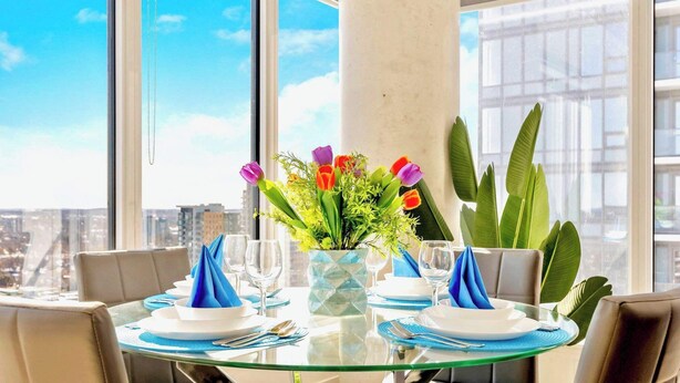 Mesa de comedor con servilletas azules, platos blancos y cristalería transparente, con vista a la ciudad, grandes ventanales y un alto pilar.