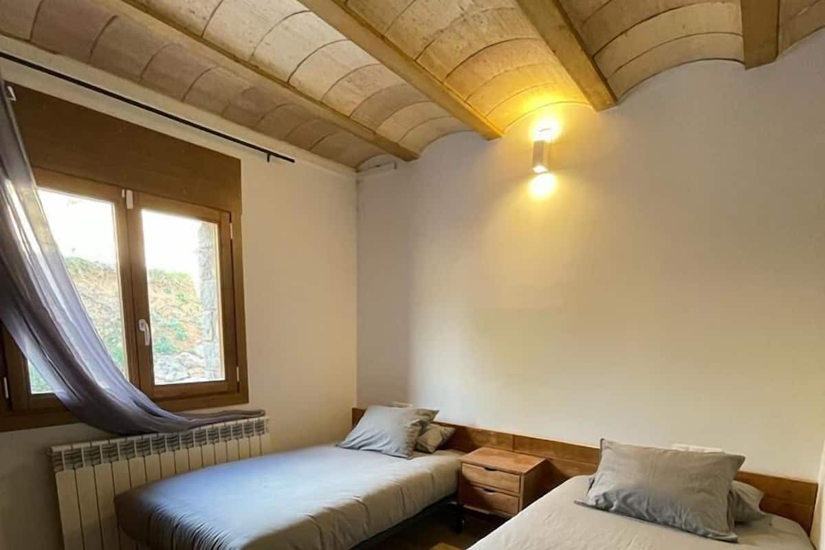 A room with two single beds, a wooden ceiling, a window with a curtain, and a wall-mounted light.