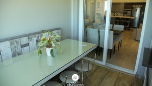 A modern kitchen with a built-in oven and refrigerator, a dining area with a glass table and chairs, and a sliding glass door leading to a living area. - Apartamento Vista Mar em Imbituba (Imbituba)