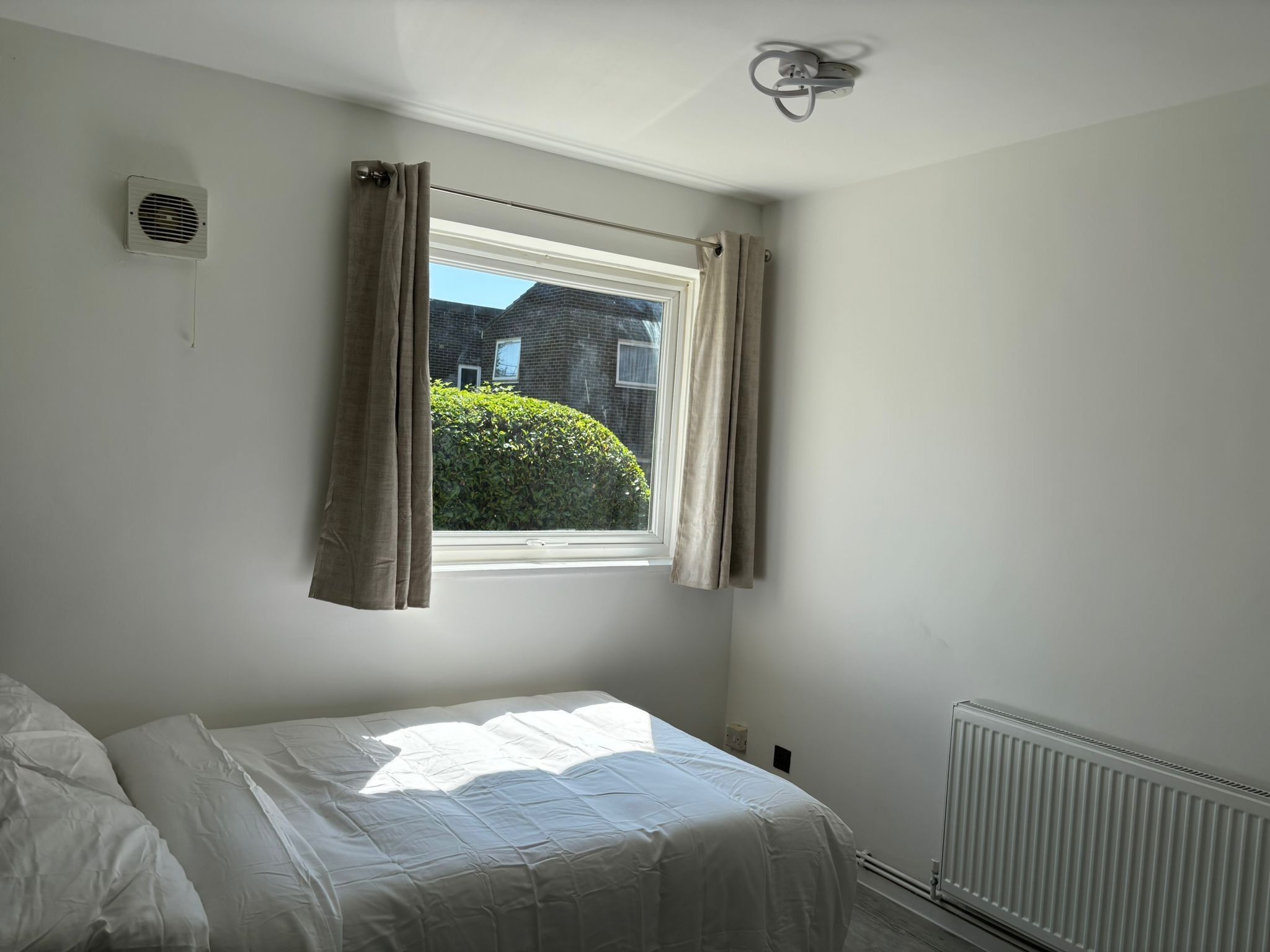 Une chambre à coucher avec un lit, une fenêtre avec des rideaux, un radiateur et un luminaire fixé au mur.