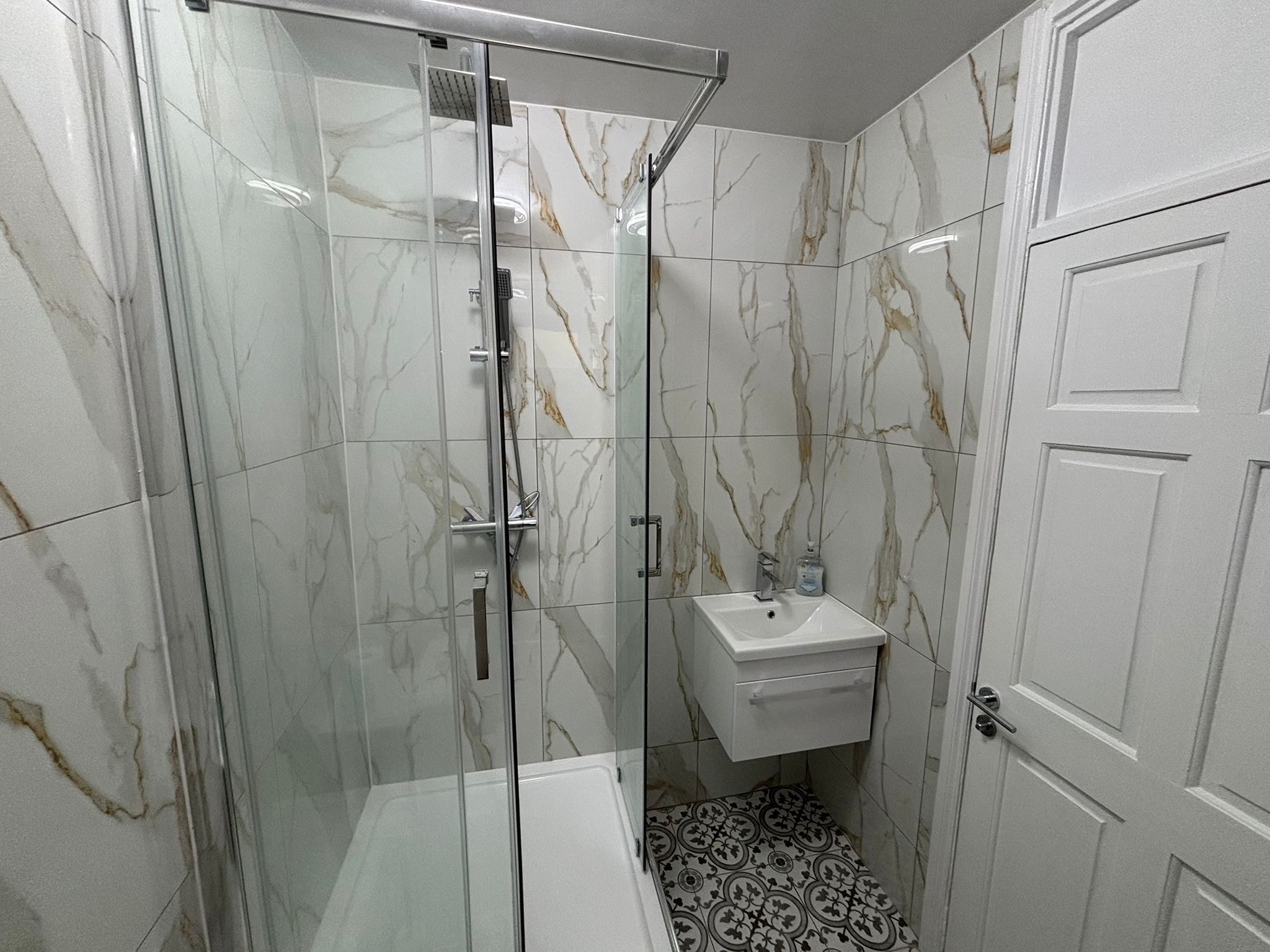 Un baño con ducha de vidrio, lavamanos blanco y baldosas de suelo con diseño.