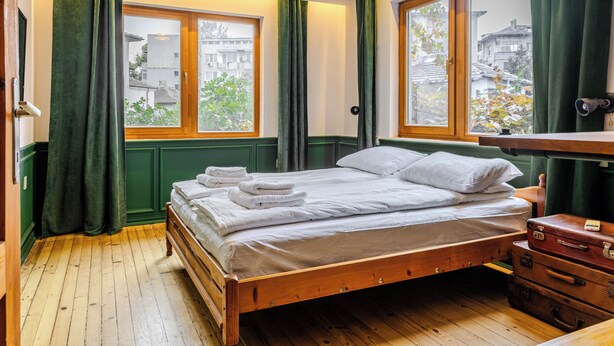 Un lit en bois, recouvert de draps blancs, avec vue sur les bâtiments à travers une fenêtre.