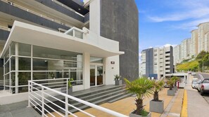 A modern building with a glass facade, a staircase leading to a flat entrance, and a parking area with cars. - Oceana Suites EuroMarina II (Viña del Mar)