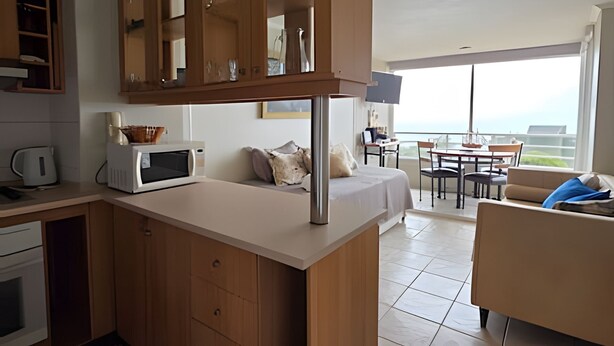 A modern kitchen with a built-in microwave, a kettle, and wooden cabinets. There is a dining area with a table and chairs, and a large window offering a view of the ocean. - Oceana Suites EuroMarina II (Viña del Mar)