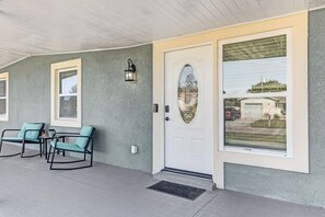 Eine überdachte Veranda mit zwei Schaukelstühlen und einem kleinen Tisch, eine weiße Tür mit dekorativem Glasfenster und ein großes Fenster mit Blick auf ein Fahrzeug und eine Landschaft.