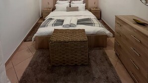 Une chambre à coucher comprenant un lit, des tables de chevet, une commode, un banc en osier et un luminaire fixé au plafond.