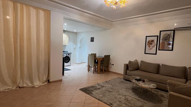 Un salon comprenant un canapé, une table basse et un tapis. On y trouve également un coin repas avec des chaises et une table. La pièce est éclairée par un luminaire au plafond et une porte donne accès à une autre pièce.