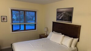 Uma cama bem arrumada com lençóis brancos, cabeceira de madeira, um quadro na parede e uma janela com persianas.