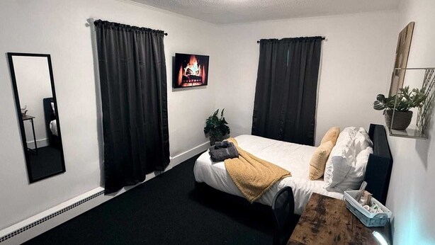 Een slaapkamer met een bed, zwarte gordijnen, een spiegel, een plant en een schilderij.