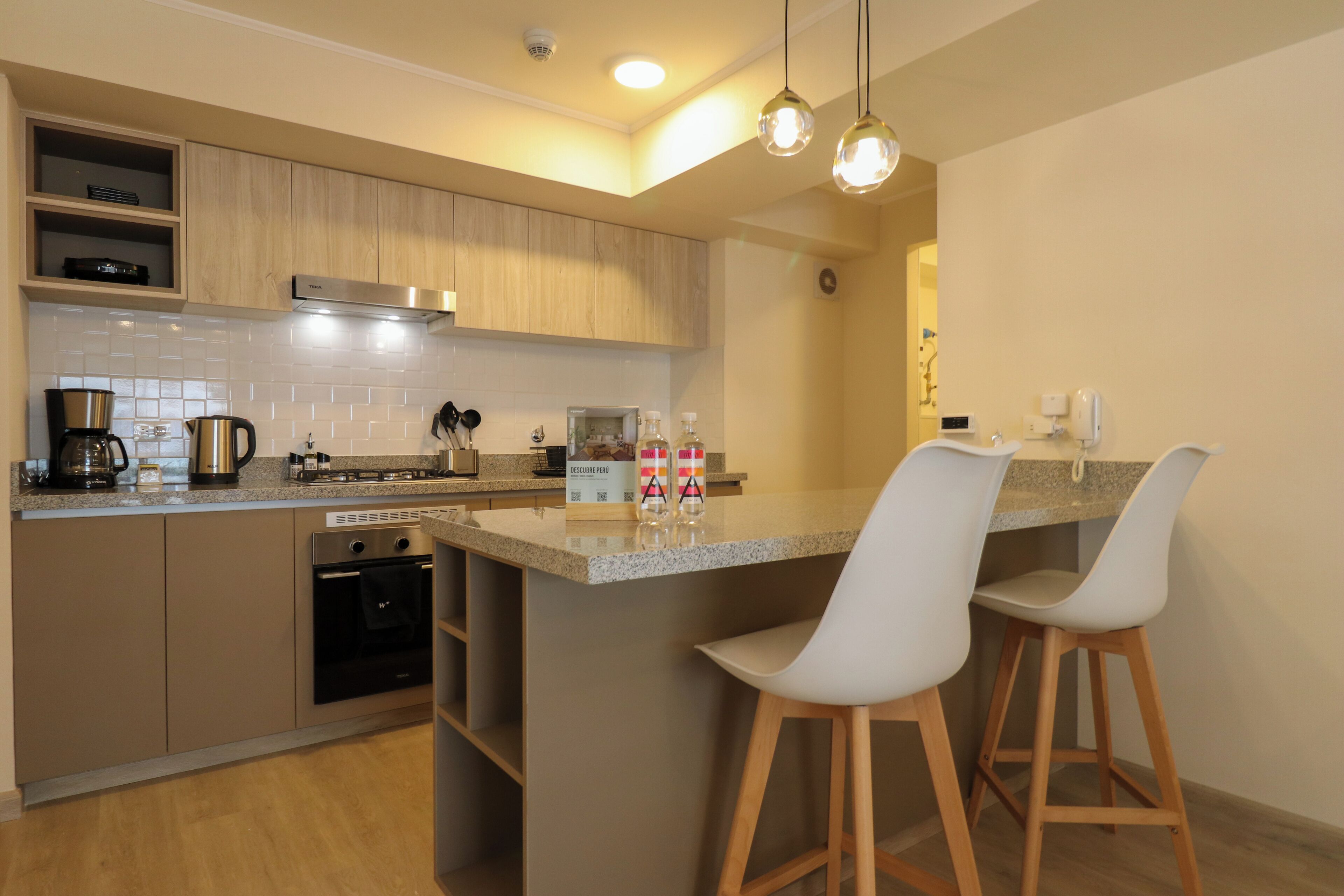 A modern kitchen with wooden cabinets, a built-in oven, a coffee maker, and two bar stools.