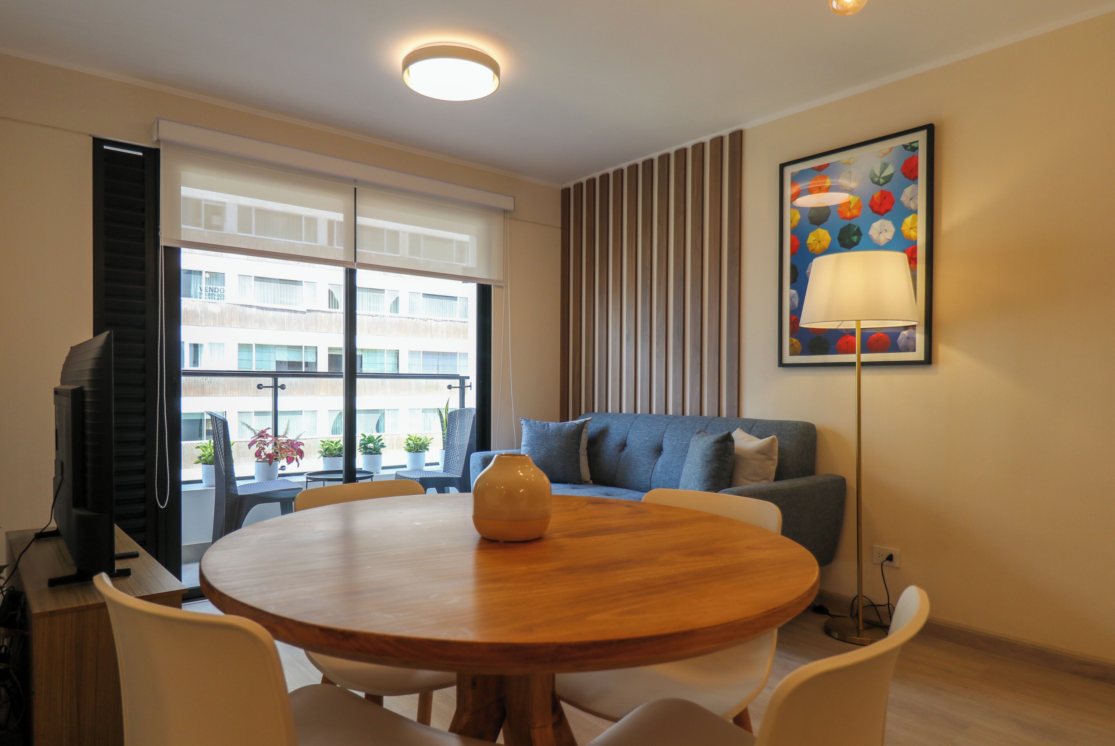 A room with a round wooden table, white chairs, a blue sofa, a floor lamp, and a painting on the wall.